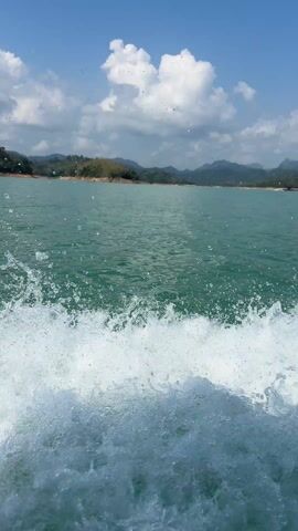 A low-angle shot shows a body of water, possibly a lake, on a sunny day. The water is a light green color, and white foamy waves are visible close to the camera, indicating movement, perhaps from a boat. The background features land with green trees and rolling hills with visible mountains in the distance, all under a blue sky with white clouds.