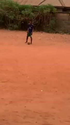 The video shows three people playing soccer on a red dirt lot. One person is wearing a blue soccer jersey and dark shorts, while the other two have no shirts and are wearing dark shorts. The person in the jersey dribbles the ball, then passes it to one of the shirtless players, who continues to dribble the ball. There is a stone wall behind the players and a building with a brown roof behind the wall. There is a person sitting down in the background to the right.