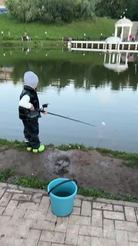 A young child wearing a grey hat, black vest, grey pants, and green boots stands on the bank of a lake, holding a fishing rod. The child is positioned facing the lake and is actively fishing. A blue bucket sits on the ground near the child. In the background, across the lake, is a grassy hill with trees and a white gazebo structure. The water reflects the surroundings, and there are a few people visible near the gazebo. The foreground shows a paved walkway with grass along the edge of the lake. The overall environment appears to be a park or recreational area.