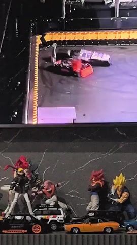 The video shows a screen displaying a robot combat event. In the foreground, a shelf holds several anime action figures and miniature cars. On the screen, two combat robots are engaged in battle. One robot is predominantly red and black, while the other appears to be dark gray or black. The robots are in motion, with sparks or debris visible, indicating active combat. The arena floor is purple and gray with yellow markings. The background of the arena is dark with orange and yellow barriers. The lighting is dramatic, with bright spotlights.