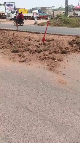 The video shows a roadside scene on a clear day. In the foreground is an asphalt road with some cracks and debris. Beyond the road is a pile of brown soil with scattered vegetation. A red shovel is stuck upright in the soil. To the left, a tree provides partial shade, under which a person wearing a yellow safety vest appears to be working near the base of the tree. In the background, across another road, there are several parked trucks, a yellow excavator, and a sign that reads "LEADGATE Construction Yard". A POS sign is also visible. A motorcyclist is seen riding on the road.