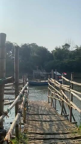 A wooden walkway made of bamboo planks leads to a body of water. The walkway is bordered by bamboo railings. A boat is docked in the water, and there is a structure on the boat. The background is filled with trees and a clear sky.