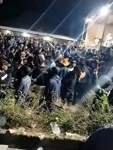 The video shows a large crowd of people gathered outdoors at night, likely for an event or performance. There is a stage set up in the background with bright lights and what appears to be performers on it. The crowd is densely packed, with many individuals visible from the waist up. Some people are holding up their hands, suggesting they are dancing or engaging with the performance. There is also evidence of vegetation and some discarded items, such as plastic cups, in the foreground. The overall atmosphere appears to be lively and energetic.