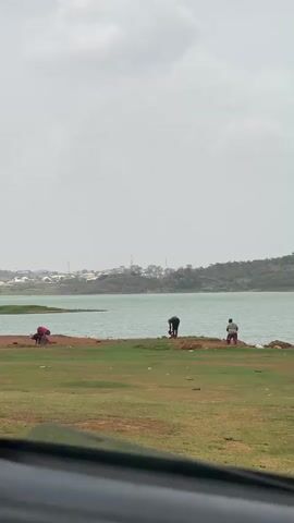 The video shows three individuals by a body of water. One person, wearing a red shirt, is crouched down near the water's edge. Another person, dressed in black, is standing and appears to be doing something with their hands near the water. The third person is walking along the shoreline, carrying a red container. The landscape includes a green grassy area in the foreground, leading to a muddy bank at the edge of the water. Across the water, there's a cityscape with buildings, trees, and hills visible in the distance under a cloudy sky. The shot seems to be taken from inside a vehicle, as part of the dashboard is visible at the bottom.