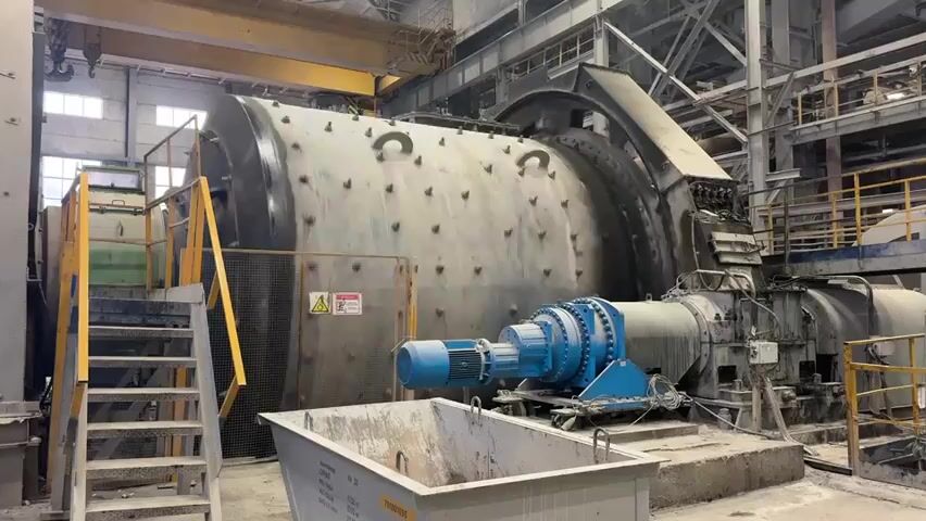 The video shows a large industrial grinding mill in a factory setting. The mill is a cylindrical drum on its side, with a blue motor and gearbox attached to one end. There are stairs leading up to a platform on the left side of the mill. A yellow crane is visible in the background. The mill appears to be dusty and worn, suggesting it is in active use. There are no people visible in the video. The environment is industrial, with concrete floors and metal structures.