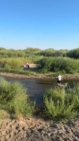 The video shows a natural scene with a river and lush greenery under a clear blue sky. In the foreground, there is dry, sandy ground with sparse vegetation. As the camera pans, a narrow river comes into view, with people on its banks. On the far bank, two individuals are seen standing near some bushes, and one appears to be holding a bag. On the near bank, a person wearing a white t-shirt and black shorts is standing on some rocks, seemingly preparing to enter or having just exited the water. Further down the river, a dark-colored vehicle is partially visible on the left side of the frame. The environment suggests a rural or wilderness setting, possibly a recreational spot for outdoor activities.