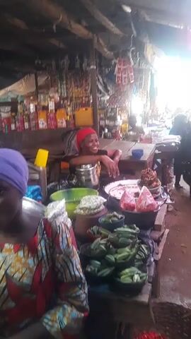 The video shows a bustling market scene, likely in an African country, filled with vendors selling various goods. The camera moves through the narrow aisles between the stalls. In the foreground, a woman with a red headscarf is seen smiling behind her vegetable stand, which displays okras, tomatoes, and other items. Other vendors sit at their tables, waiting for customers. One woman in a black robe passes by. The stalls are under a corrugated metal roof that lets in sunlight. The tables are cluttered with produce, bowls, and other objects.