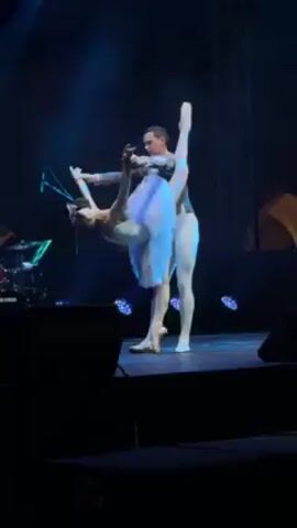 On a dark stage, two ballet dancers perform a partnered routine. The female dancer is en pointe, wearing a white tutu and tiara, while the male dancer is in a short, decorative jacket and tights. They are in a pose, with the male dancer holding the female dancer as she extends her leg high in the air. The stage is lit with blue lights and there is some musical equipment visible in the background.