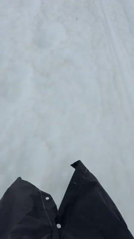 The video shows a first-person perspective of someone walking or skiing on a snow-covered path. The path is marked by tire tracks, suggesting it might be a road or a trail used by vehicles. The terrain is mostly flat with a slight incline, leading towards a mountainous landscape. The sky is overcast, creating a dim and moody atmosphere. In the distance, two figures are visible, appearing to be walking or skiing in the same direction. The snow is somewhat dirty, with patches of brown and gray visible, indicating it may not be freshly fallen. The overall environment appears to be a remote, natural setting, possibly a mountain range or a highland area.