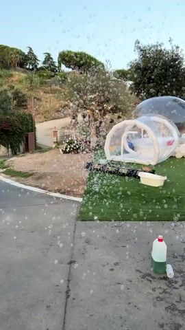 Bubbles fill the foreground and mid-ground of the video. In the mid-ground, a large inflatable bubble structure is set up on a patch of artificial grass. A white tub is to the right of the inflatable. To the left of the inflatable are rows of black objects that appear to be bubble machines. In the background, a dirt path leads up a gentle slope, flanked by trees and foliage. A white fence and white flowers are visible on the left side of the path. In the far right of the mid-ground, a large clear bubble structure is visible. In the bottom right corner of the frame, a large white plastic bottle with a red cap and a smaller clear plastic bottle are on the ground.