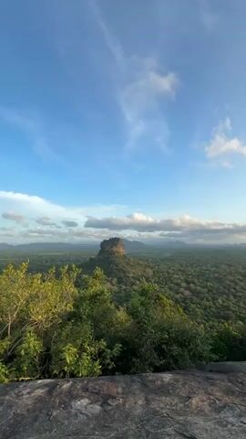 The video shows a landscape with a large rock formation in the distance, surrounded by lush green forest under a blue sky with scattered clouds. The camera pans slowly from left to right, revealing more of the forest and sky. The foreground consists of a rocky surface, which appears to be the edge of the hill where the video is taken from.