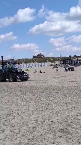 A blue tractor drives across a sandy beach on a sunny day with a partly cloudy sky. In the background are buildings and people on the beach. The tractor is labeled "NEW HOLLAND". The beach appears to be crowded in the distance. The camera follows the tractor as it moves across the sand.