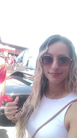The video is a first-person perspective shot of a woman wearing sunglasses, a white top, and jewelry. She stands in a crowded outdoor area. The background shows many people, some wearing red shirts. There are tents and vehicles in the background. The camera moves slightly, capturing different people and objects as the woman adjusts her hair and smiles. Overall, the video depicts a lively outdoor event with a crowd of people.