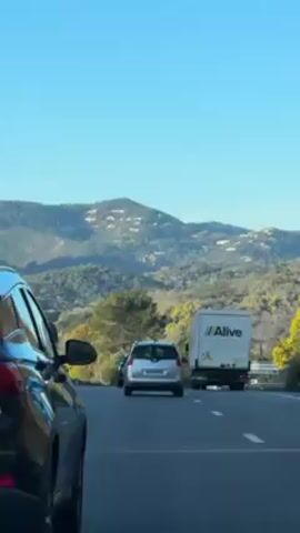 The video depicts a view from a car driving on a highway, with other cars and a truck visible on the road. The backdrop is a mountain range under a clear blue sky. The landscape includes trees and other vegetation. The car from which the video is taken has a license plate that reads "GP-659-GF".  The truck displays the word "Alive" in its logo.
