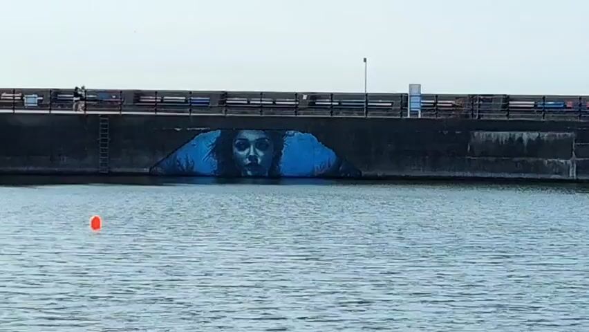 The video shows a large mural of a woman's face painted on a concrete wall that is partially submerged in water. The mural is predominantly blue and white, with the woman's eyes looking directly at the camera. The water in front of the wall is calm and reflects the sky. There is an orange buoy floating in the water to the left of the mural. In the background, there is a walkway on top of the wall with several people walking and sitting on benches. The sky is overcast, and the overall lighting suggests it is daytime. The mural appears to be part of a larger urban landscape, possibly a harbor or waterfront area.