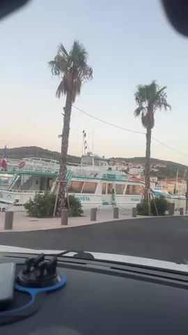 The video begins with a view from inside a vehicle, looking out at a waterfront scene. A white boat with blue trim is docked on the left side of the frame, with the name "Aquavista" visible on its side. Two tall palm trees are situated in front of the boat. The boat is large and appears to be a passenger vessel. Beyond the boat and palm trees, a town with buildings and a marina can be seen in the background. The sky is a pale blue, suggesting either early morning or late evening. The camera pans slightly to the right, revealing more of the marina with several other boats docked. The town's architecture is visible, with buildings clustered on a hillside. Another palm tree is visible on the right side of the frame. The foreground of the video shows a portion of the vehicle's dashboard, including a phone mounted on a suction cup holder. The overall impression is a scenic view of a coastal town and harbor.