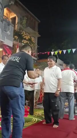 In an outdoor setting at night, likely a celebration or event, a man in a black t-shirt with "OFFICIAL" printed on the back is facing a man in a white polo shirt and black pants. The man in black is gesturing with his right hand towards the man in white, who appears to be looking at him and smiling. Several other men are visible in the background, some in white shirts and others in different attire. Decorative flags are strung across the background. A red carpet is laid out on the ground, with a patterned rug underneath. A building is visible behind the group, with foliage growing on it. The overall atmosphere seems lighthearted, indicated by the smiles on some of the faces.