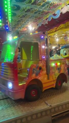 The video shows a children's carnival ride at night. The ride consists of various colorful vehicles, including what appears to be a truck, cars designed to look like cartoon characters such as Donald Duck and a Dalmatian, and a bug-shaped car. These vehicles are moving along a track, and many have bright, flashing lights. People, including children and adults, are visible in and around the vehicles, some appearing to be riding the attraction. The overall environment is festive and illuminated by numerous lights.
