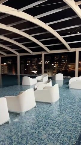 This is a video of an empty swimming pool at night. The pool is covered by a canopy with a white frame and dark panels. White lounge chairs are partially submerged in the water. In the background, city lights are visible. The camera pans slowly across the pool, showing the chairs and the surrounding environment. There are no people or animals in the video.