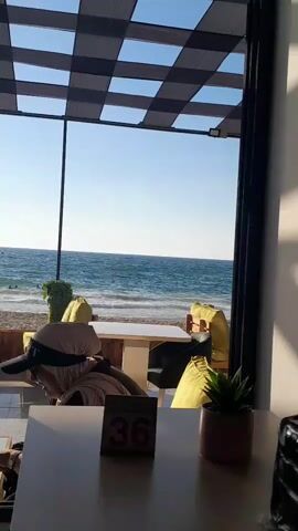 The video shows a view from inside a building, looking out at a beach and the ocean. A checkered awning is visible overhead. In the foreground, there is a table with a small potted plant and a table number. A person wearing a hijab and a hat is seated in the middle ground, facing away from the camera and towards the beach. The beach is visible with people in the water and on the sand. The ocean is calm with small waves. The sun is shining, creating shadows on the floor inside the building.