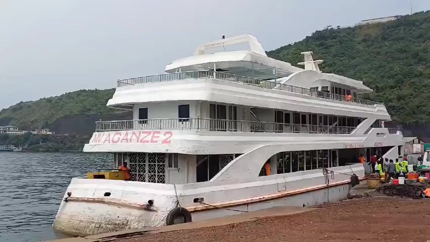 The video shows a white, multi-decked boat named "MV AGANZE 2" docked at a pier. The boat has multiple levels with windows and railings, and some people are visible on board and along the pier. There are also some vehicles and containers nearby, along with greenery on the slope of a hill visible in the background. The overall environment suggests a port or harbor setting, possibly preparing for a trip.
