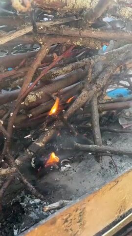 The video shows a close-up view of a small fire burning among a pile of dry branches and twigs. Flames are visible, flickering and consuming the wood. Smoke rises from the fire, obscuring some of the branches. The fire appears to be contained within a metal structure or pit, with the edge of what looks like a wooden border visible in the foreground. The overall impression is of a controlled burn of natural materials.