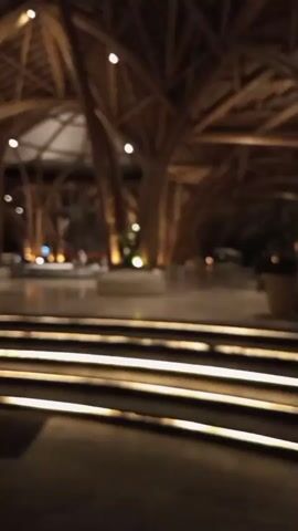 A blurred video shows the interior of a building with a high, arched ceiling constructed from bamboo. The space is dimly lit, with warm lights illuminating the bamboo structures and the floor. In the foreground, there are illuminated steps. In the background, there appear to be seating areas and plants. The overall impression is of a modern, possibly upscale, indoor space with a naturalistic architectural design.