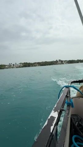 The video shows a view from the back of a boat moving through turquoise water. The camera is positioned facing forward and slightly to the right, capturing the wake of the boat and the distant shoreline. The shoreline features lush green trees and several white buildings, possibly houses or resorts. The sky is overcast with gray clouds. A blue rope is visible in the foreground, attached to the railing of the boat. The railing itself appears weathered. The overall impression is a travel or scenic view from a boat on a cloudy day.