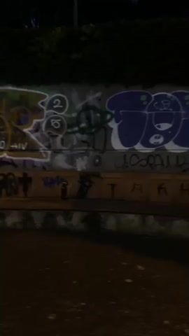 This video captures a series of dimly lit urban scenes, likely at dusk or night, in Logroño, Spain. The initial frames show a graffiti-covered wall, with various tags and artistic pieces in spray paint. Visible text includes "CALERO," "HIV," and "TARPA." The wall appears to be part of a structure with a curved concrete base, suggesting it might be a retaining wall or part of an amphitheater-like setting. Bushes and trees line the top of the wall, and a street lamp illuminates a section of the path alongside it.

The video then transitions to a person walking across a bridge. The bridge has red railings and a yellow pathway. The person, viewed from behind, is dressed in dark clothing and appears to be carrying a bag. The sky is a deep blue, indicating the evening is well underway. The surrounding landscape is dark, with silhouettes of trees visible against the twilight sky. A body of water, possibly the Ebro River, can be seen below the bridge. The overall mood is quiet and solitary, with the urban environment illuminated by artificial lights.