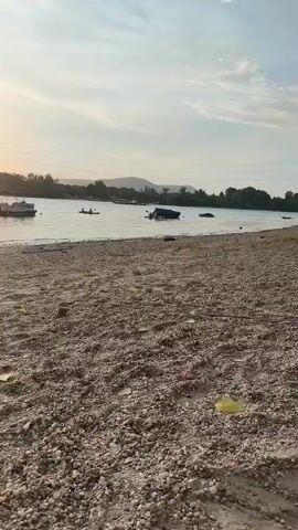 This video captures a serene sunset scene at a beach in Dunakeszi, Hungary. The foreground is dominated by a sandy and pebbly shore, scattered with a few debris items, including a single yellow plastic bag and a pair of black sandals.

In the middle ground, the calm water of a lake or river reflects the warm, golden hues of the setting sun, which is partially obscured by a distant transmission tower. Several boats are visible on the water: a pontoon boat with a canopy, a smaller boat, and what appears to be a capsized or submerged vessel. Two individuals are seen in the water, one on a kayak and another person, presumably a man, standing with a dog near the shoreline.

The overall atmosphere is peaceful and tranquil, suggesting a leisurely end to the day. The time of day is clearly indicated by the low position of the sun and the soft, diffused light. The presence of the transmission tower and the type of vegetation on the distant shore provide subtle clues about the geographical location. There is no visible text in the video.