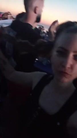 A woman is filming herself with a phone at an outdoor event. She has dark hair pulled back in a ponytail with some strands loose framing her face. She is wearing a black top with straps. Her eyes are lined with dark makeup, and she has a small sparkling accent near her left eye. Her skin appears flushed pink from lighting. In the background, other people are visible, some in focus and some blurred. A man with short, dark hair and a beard is visible over her left shoulder. The sky behind the people suggests it is dusk, with a soft gradient of pink and blue. Some of the people in the background appear to be standing and interacting, perhaps at tables or a gathering. The lighting shifts from pink to red throughout the clip.