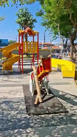 The video shows a playground with a red and yellow slide structure. In the foreground, there is a spring rider, possibly a horse, that appears to be made of bamboo and metal. The rider is secured with ropes and is on a black mat. The playground is paved with gray and tan tiles, and there are shadows from trees. In the background, there are trees, buildings, and a blue sky.