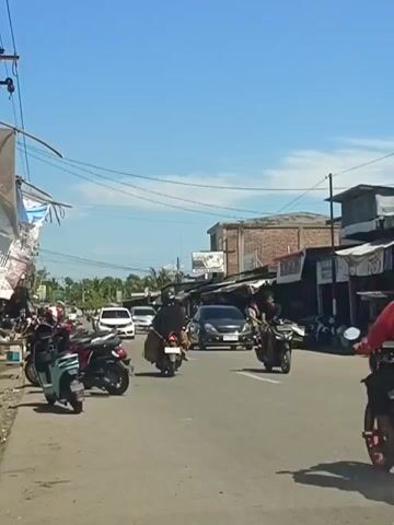 The video shows a street scene with several motorbikes and cars. In the foreground, there are two motorbikes parked on the left side of the road. A light blue motorbike is on the left, and a red motorbike is next to it. Further down the road, a person in a red jacket is riding a motorbike. To the right, there are several shops with awnings. A dark-colored car is driving in the middle of the road. More motorbikes are seen on the right side of the road. The sky is clear and blue. The scene appears to be in a developing area with some vegetation visible in the background.