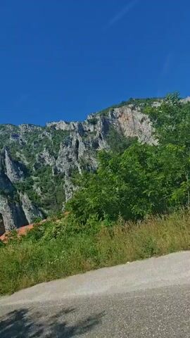 The video pans across a picturesque landscape under a clear blue sky. The scene is dominated by a dramatic mountain range with rugged, rocky cliffs dotted with green vegetation. In the foreground, a lush green hillside slopes down to a paved road or path. Scattered amongst the trees are glimpses of buildings with terracotta roofs, suggesting a village or settlement nestled into the mountainside. The bright sunlight indicates it is daytime, likely a warm and pleasant day. The overall impression is one of natural beauty and tranquility, with a hint of human habitation integrated into the wild landscape. The camera movement suggests it is being filmed from a moving vehicle, perhaps a car, as it travels along the road. There is no visible text in the video. The location is identified as Lyutibrod, Bulgaria, which aligns with the mountainous terrain and architectural style hinted at in the footage.