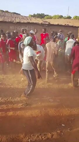 The video shows a group of people in an open area, kicking up dust. Many are wearing red jerseys, and one woman is wearing a light blue headscarf. At one point, a person wearing a large mask and a grey robe moves into the foreground. The area appears to be in or near a village or rural setting, with buildings visible in the background. The atmosphere seems festive, with people moving around and creating dust as they walk and possibly dance.