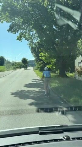 A person on a bicycle rides down a paved road on a sunny day. The cyclist is wearing a light blue shirt, grey shorts, and a helmet. The road is bordered by trees and grass on the right side. In the background, there are more trees and a clear blue sky with contrails from airplanes. The video appears to be filmed from a vehicle, as indicated by the dashboard visible at the bottom of the frame.