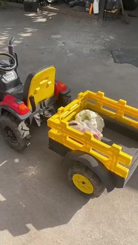 The video shows a stationary, bright red and black toy tractor with a yellow seat, attached to a yellow toy trailer. The trailer contains what appears to be white and green floral arrangements. The scene is outdoors, with a concrete or asphalt ground surface. Sunlight casts shadows on the ground, indicating daytime. The tractor has large, rugged tires and a steering wheel. The trailer has a sturdy yellow frame and wheels. The background is slightly blurred, but appears to be an outdoor setting with some vegetation and possibly structures.