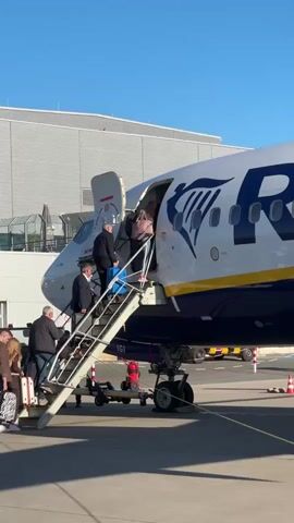 The video captures an outdoor scene at an airport, likely in Weeze, Germany, under a bright, clear blue sky. A large, white and blue Ryanair airplane is prominently featured, parked on the tarmac. Several passengers are actively boarding the aircraft via a set of mobile boarding stairs positioned at the front left entrance.

Individuals, including men and women, are seen ascending the stairs. Some are carrying luggage, such as small wheeled suitcases and handbags, as they make their way into the plane. Near the entrance, a person in a dark uniform, possibly a pilot or ground crew, is also visible. The boarding stairs have the identifier "1G1" printed on their side.

In the background, a modern airport terminal building with a grey facade is visible, suggesting the plane is parked at a gate or stand. The weather appears sunny and clear, indicating fair conditions for air travel. The activity depicted is a routine passenger boarding process for a Ryanair flight.