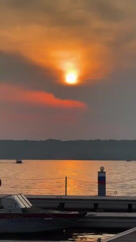 A sunset over a body of water. The sun is partially obscured by clouds, casting a warm orange glow. In the foreground, a dock with several boats is visible. One boat in the foreground has the markings "TA3092 RUS 50" on its side. In the distance, several small boats are moving across the water. The horizon is lined with trees.