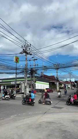 A busy intersection features multiple motorbikes navigating the roadway under a cloudy sky. Overhead, a dense network of power lines crisscrosses the frame, supported by utility poles. To the left, a convenience store with green and white signage is visible, along with parked vehicles and some pedestrians. The road surface is asphalt. Motorcyclists in casual attire are the primary figures, engaged in the act of riding. The overall scene suggests a typical urban or suburban environment in a region where motorbike transportation is common.