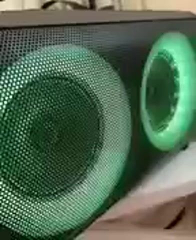 The video shows a close-up, side-view of a portable speaker. The speaker is black with a mesh grille covering two circular openings, presumably for the speakers. The openings have illuminated rings around them, which change color throughout the short clip. The first color shown is green, followed by white, then pink. The speaker appears to be resting on a surface covered with a light-colored fabric. The lighting in the environment is dim, highlighting the illuminated rings on the speaker. No people are visible in the video. The camera angle is static, and the focus is on the speaker itself. The video is very short, only four seconds long, and appears to be a demonstration of the speaker's lighting features.