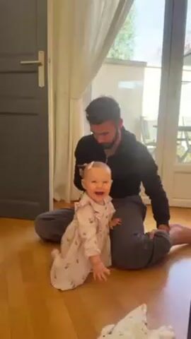 A man and a baby are indoors on a wood floor. The man is sitting on the floor with his legs crossed, wearing a black shirt and grey pants. The baby is kneeling, wearing a patterned dress with a pink bow in her hair. She initially smiles broadly, then opens her mouth wide as if laughing or crying. The camera zooms in closely on the baby's face during this action. Behind them are sliding glass doors leading to an outdoor patio with chairs and tables, partially obscured by a white curtain. A grey door is to the left. A small toy or cloth is on the floor near the bottom of the frame. The lighting is bright and natural.