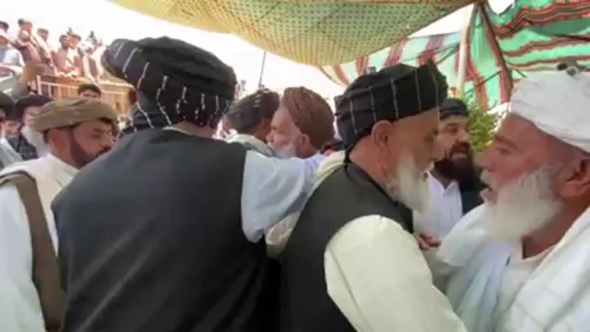 Here's a detailed description of the video:

**Scene:**

The video appears to show a gathering of men outdoors, likely in a rural or semi-rural setting. They are standing under a tent-like structure made of fabric stretched overhead, possibly to provide shade.

**People:**

The majority of the people in the video are men, most of whom appear to be older. Many of them have beards. They are wearing traditional clothing, including turbans of various colors (white, black with patterns, brown). Many are wearing vests over light-colored, long-sleeved shirts.

**Actions and Interactions:**

*   The men are greeting each other.
*   There's interaction and close physical contact, including hugging.
*   People are talking to each other, with some gestures.
*   Someone is filming with a mobile device.

**Environment:**

*   The environment is outdoors and appears to be a dusty place.
*   The tent-like structure provides shade.
*   In the background, there are other people and some undefined objects.

**Overall Impression:**

The scene seems to depict a social gathering of men, potentially a celebration, meeting, or a gathering of elders. The clothing suggests a cultural or regional context that is likely South Asian or Middle Eastern.