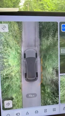 The video shows a car's internal display screen. The top portion of the screen displays a 3D bird's-eye view of the car driving down a narrow paved road bordered by dense green foliage on both sides. The car's tires are positioned on the right side of the road. A distance of 87 cm is shown on the screen, indicating the car's proximity to an object on its right. As the video progresses, the car moves forward, and the displayed distance decreases to 67 cm and then to 15 cm. The camera view then shifts to show a real-time feed from the front of the car, displaying the road ahead, which is also bordered by trees and foliage. A sign with a speed bump symbol is visible on the right side of the road in the distance. The bottom portion of the screen displays additional camera views and control icons, including a rear-view camera and options for selecting different camera perspectives. The scene appears to be a rural or suburban environment.