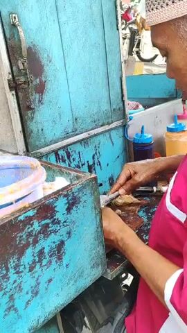 The video shows a close-up view of a street food vendor preparing food. The vendor, whose face is partially obscured, is wearing a pink apron and a patterned head covering. They are using a knife to cut or prepare food items on a wooden surface. In the foreground, there is a blue wooden cart with various containers. One container is a large, clear plastic tub with a purple lid, filled with a light brown liquid and a wooden utensil. Another metal pot contains what appears to be fried food items. To the right, there are two squeeze bottles, one with a blue cap and the other with an orange cap, presumably containing sauces. The background suggests an outdoor setting with a concrete wall and a motorcycle visible. The overall scene depicts a busy, everyday street food preparation environment.