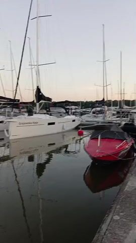 The video shows a marina with several sailboats docked. The water is calm and reflects the sailboats and their masts. There is a red dinghy in the foreground. The sky is overcast. The video pans slowly across the marina.
