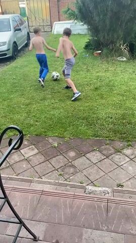 Two young boys, shirtless, play soccer in a grassy backyard. One boy wears blue pants and the other grey shorts. A soccer ball is visible on the grass. The yard has a paved patio area in the foreground and a fence and a car in the background.