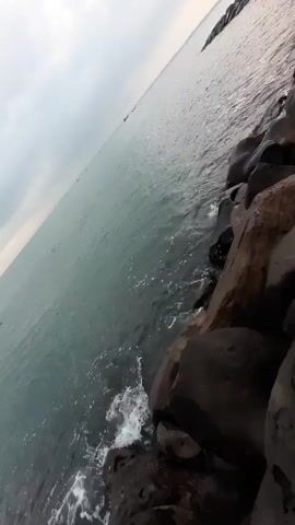 The camera is positioned at an extreme angle overlooking the sea and a rocky shoreline. The water is calm, with small waves gently lapping against the dark, irregularly shaped rocks. The sky is overcast, with a pale grey color suggesting an overcast day. In the distance, faint outlines of boats can be seen on the horizon. The scene appears to be a tranquil coastal environment, with the focus on the natural elements of the sea and rocks.