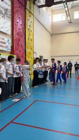 The video shows a group of teenagers standing in a line in a gymnasium. They are receiving medals and certificates from two individuals dressed in blue and red athletic uniforms. Most of the teenagers are wearing white t-shirts with matching graphics and writing, and jeans or casual pants. A girl with long hair walks by, carrying a handful of yellow ribbons, presumably for the medals. The gymnasium has a blue floor with red lines and a climbing wall on the left. Several adults stand nearby, observing and clapping. There are cones set up near the right side of the frame.