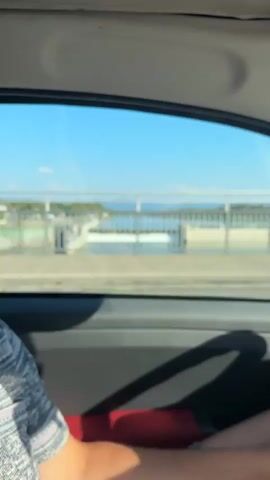 The video shows a view from inside a vehicle, looking out the side window. The camera is moving, suggesting the vehicle is in motion. Through the window, a dam or weir is visible, with water flowing over it, creating a cascading effect. Beyond the dam, a body of water stretches out, meeting the horizon under a clear blue sky with scattered clouds. The surroundings appear to be natural, with trees and hills in the distance. There are also structures that look like buildings and a radio tower in the background. In the foreground, through the car window, another vehicle is partially visible, and what appears to be the armrest and part of a person's leg are in the lower portion of the frame. The lighting suggests it is daytime, likely sunny.